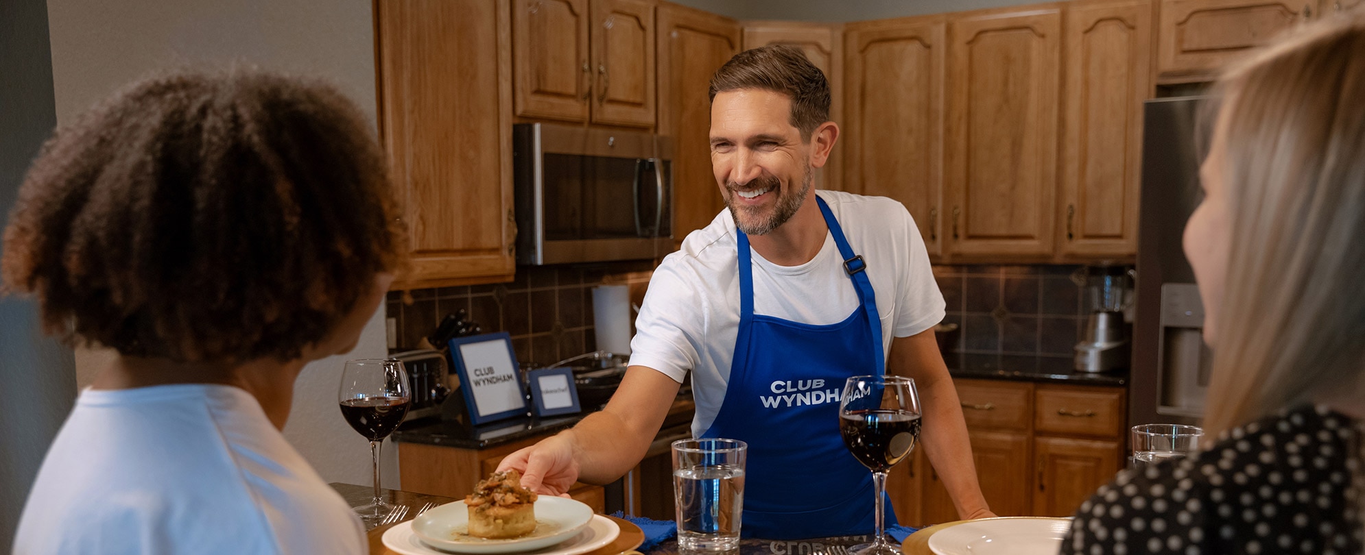 Private chef handing a plated meal to two guests in the kitchen of their Club Wyndham resort suite.