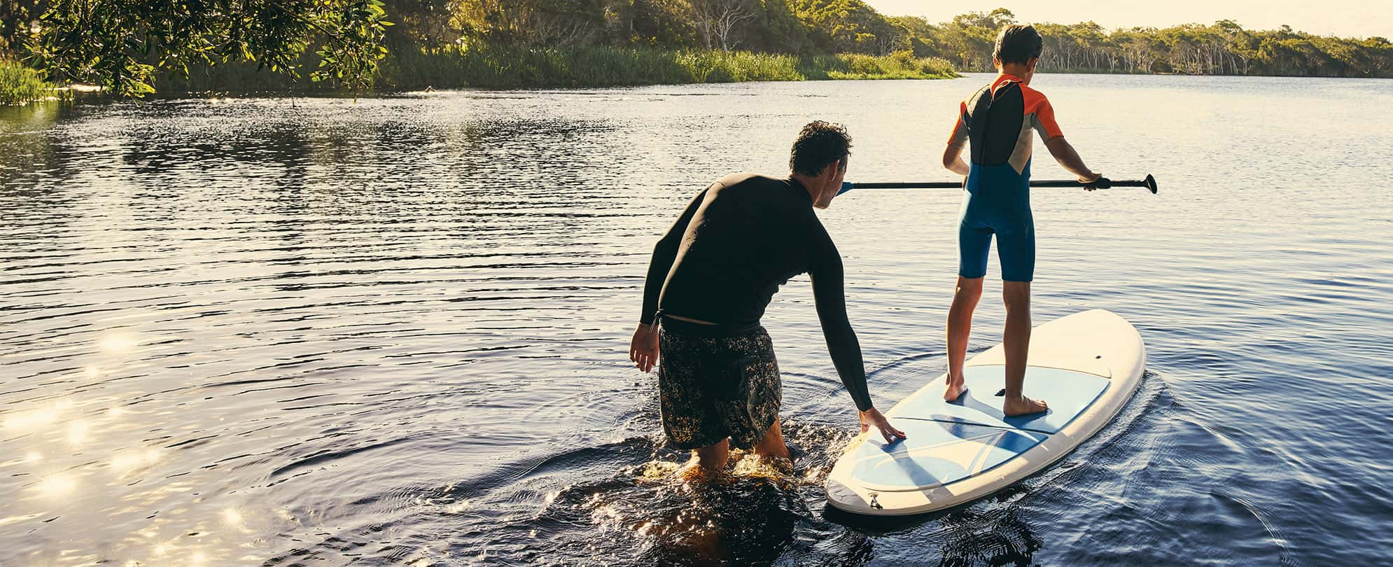 A father walks in the water behind his son who is holding a paddle on a standup paddleboard.